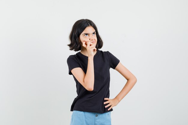 Little girl in black t-shirt, shorts standing in thinking pose and looking hesitant , front view.