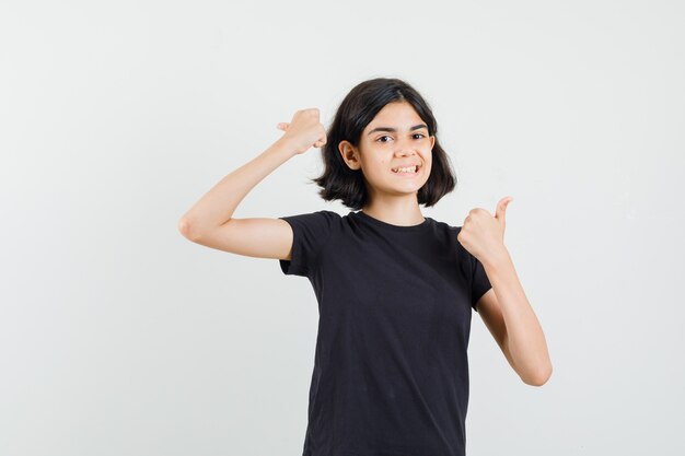 Маленькая девочка в черной футболке показывает двойные пальцы вверх и выглядит весело, вид спереди.