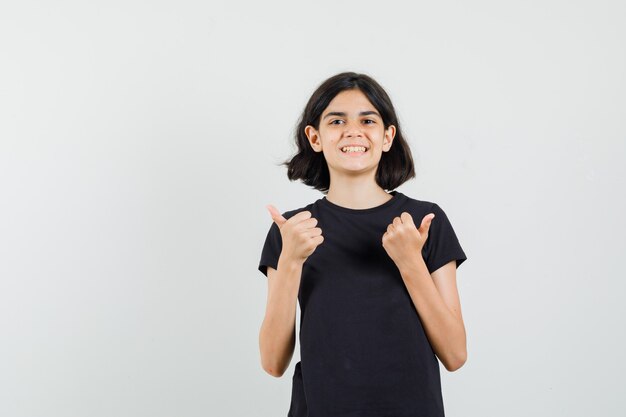 Маленькая девочка в черной футболке показывает двойные пальцы вверх и выглядит счастливой, вид спереди.