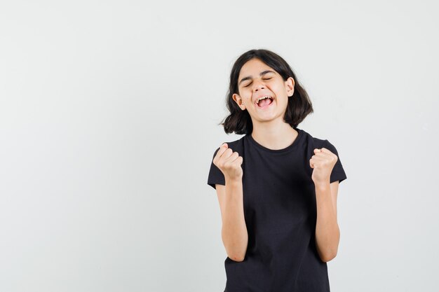 Маленькая девочка в черной футболке показывает жест победителя и выглядит счастливым, вид спереди.