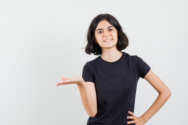 Little girl in black t-shirt spreading palm aside and looking cheerful , front view.