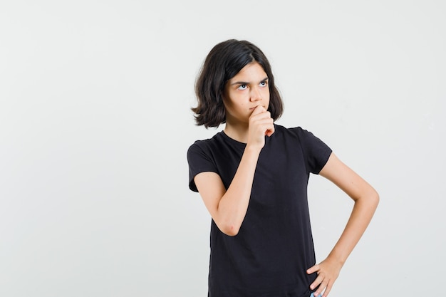 Little girl in black t-shirt standing in thinking pose and looking sensible , front view.