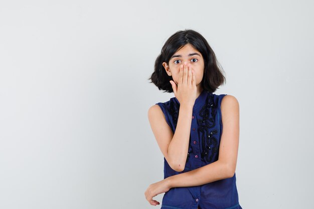 Little girl in blue blouse holding hand on mouth and looking surprised , front view.