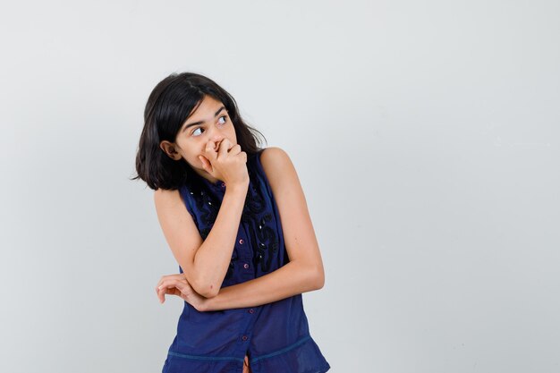 Little girl in blue blouse holding hand on mouth, looking up and looking curious , front view.