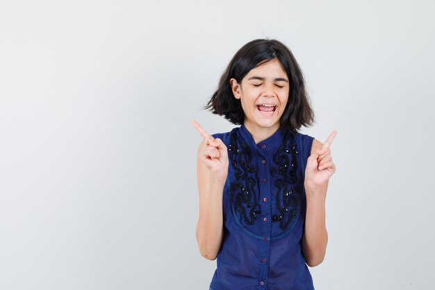 Little girl in blue blouse pointing fingers up and looking blissful