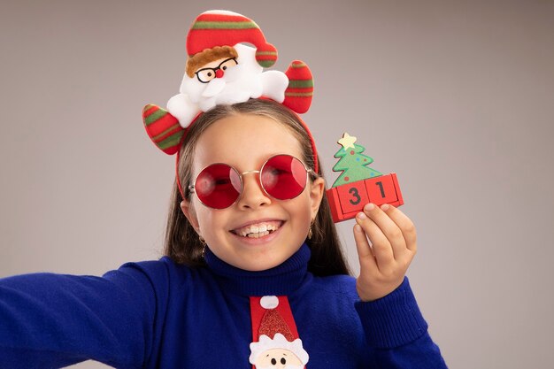Маленькая девочка в синей водолазке в забавной рождественской оправе на голове держит игрушечные кубики с новогодней датой, счастливая и взволнованная, стоя над белой стеной