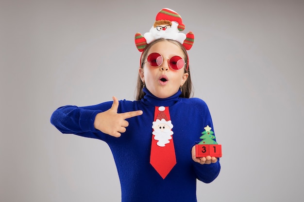 Little girl in blue turtleneck wearing funny christmas rim on head holding toy cubes with happy new year date  looking surprised pointing with index finger at cubes standing over white background