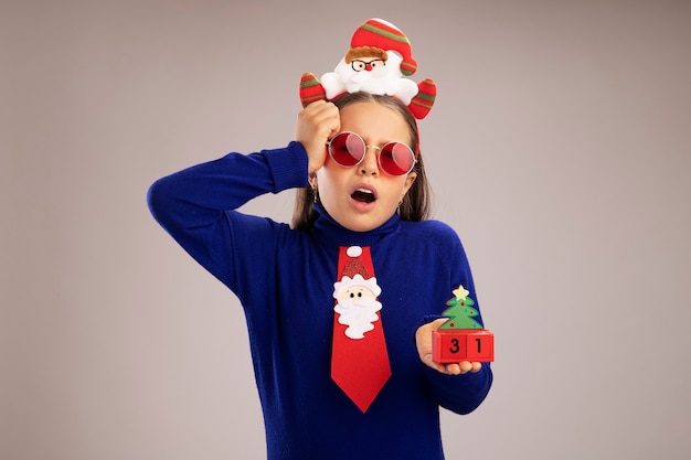 Маленькая девочка в синей водолазке в забавной рождественской оправе на голове держит игрушечные кубики с новогодней датой, удивленная и смущенная, стоя над белой стеной