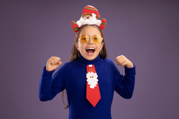 赤いネクタイと頭に面白いクリスマスの縁を持つ青いタートルネックの少女は、紫色の背景の上に立って幸せで興奮した叫び声を握りしめ拳を握り締めます