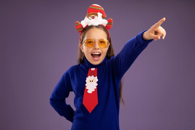Маленькая девочка в синей водолазке с красным галстуком и забавной рождественской оправой на голове счастлива и взволнована, глядя в камеру, указывая указательным пальцем в сторону, стоя на фиолетовом фоне
