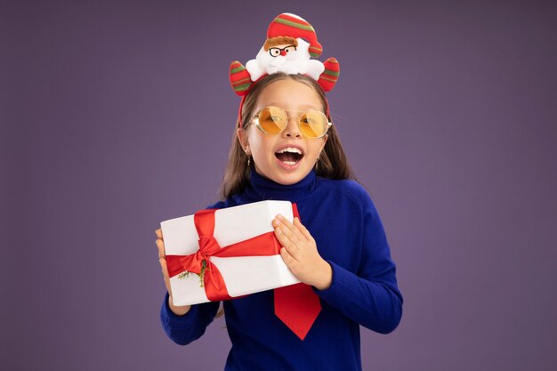 Маленькая девочка в синей водолазке с красным галстуком и забавной рождественской оправой на голове держит подарок с улыбкой на лице, счастливая и веселая, стоя над фиолетовой стеной