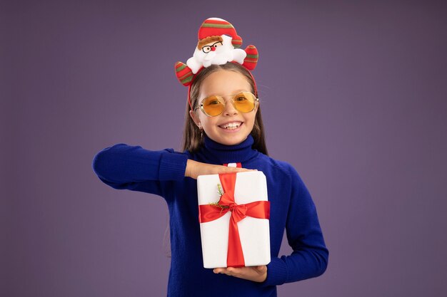 Little girl in blue turtleneck with red tie and  funny christmas rim on head holding a present   with smile on face happy and cheerful standing over purple wall