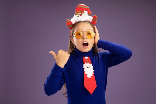 Little girl in blue turtleneck with red tie and  funny christmas rim on head looking at camera amazed pointing with thumb to the side  standing over purple background