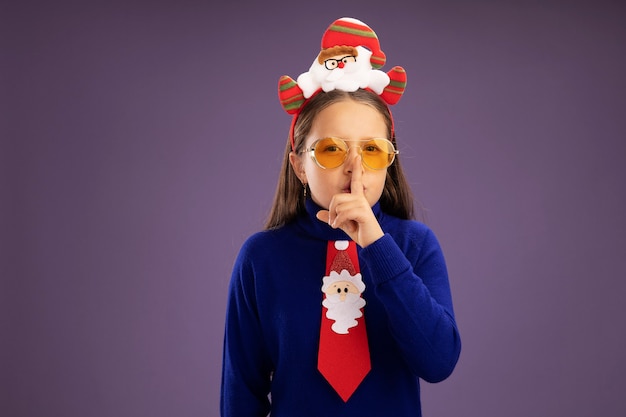 보라색 배경 위에 서있는 입술에 손가락으로 침묵 제스처를 만드는 카메라를보고 머리에 빨간 넥타이와 재미있는 크리스마스 테두리와 파란색 터틀넥에 어린 소녀