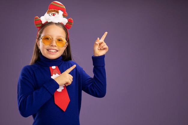 Little girl in blue turtleneck with red tie and  funny christmas rim on head looking at camera smiling cheerfully pointing with index fingers to the side  standing over purple background