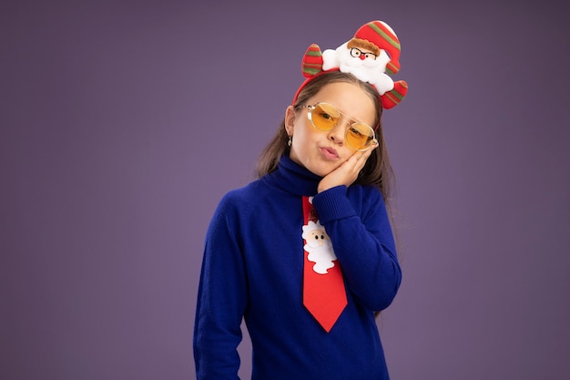 Little girl in blue turtleneck with red tie and  funny christmas rim on head looking at camera tired and bored  standing over purple background