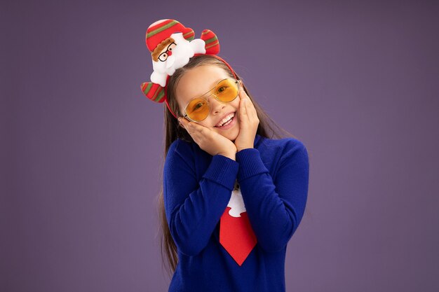 Little girl in blue turtleneck with red tie and  funny christmas rim on head looking at camera with happy face smiling cheerfully standing over purple background