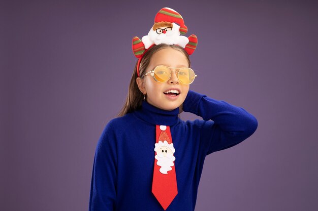 빨간 넥타이와 보라색 배경 위에 유쾌하게 서있는 행복한 얼굴로 카메라를보고 머리에 재미있는 크리스마스 테두리와 파란색 터틀넥에 어린 소녀