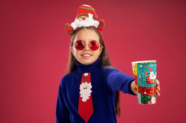 빨간 넥타이와 다채로운 종이 컵 행복하고 긍정적 인 유쾌하게 웃는 머리에 재미있는 크리스마스 테두리와 파란색 터틀넥에 어린 소녀