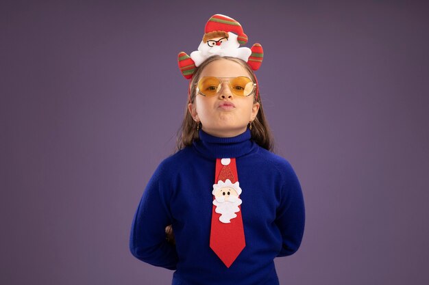 Маленькая девочка в синей водолазке с красным галстуком и забавной рождественской оправой на голове с уверенным выражением лица стоит над фиолетовой стеной