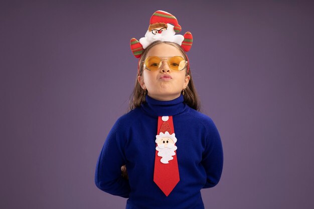 Little girl in blue turtleneck with red tie and  funny christmas rim on head  with confident expression  standing over purple wall