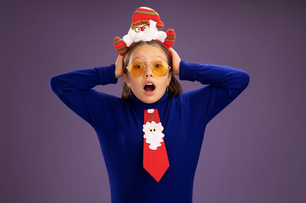 빨간 넥타이와 머리에 재미있는 크리스마스 테두리와 파란색 터틀넥에 어린 소녀 걱정하고 그녀의 머리에 손을 놀라게