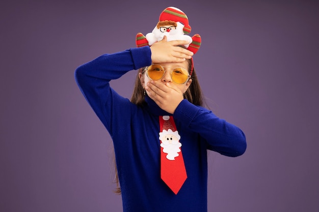 赤いネクタイと頭に面白いクリスマスの縁を持つ青いタートルネックの少女は彼女の額とcに手で心配している