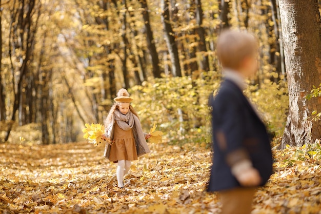 Little girl and boy in autumn park. Girl holding a yellow leaves and walking. Boy is blurred on the photo.