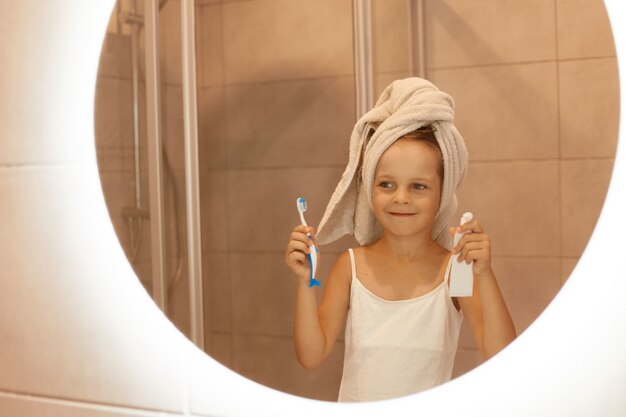 Little girl brushing teeth in bathroom, looking at her reflection in the mirror, wearing white t shirt and wrapped her hair in towel, holding toothbrush and toothpaste in hands.