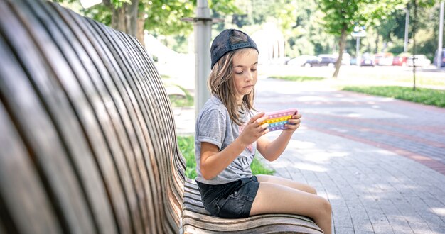 모자를 쓴 어린 소녀는 여름날 공원 벤치에 앉아 스마트폰을 사용합니다.
