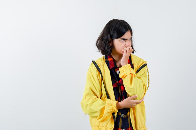 Little girl in checked shirt, jacket holding hand on mouth and looking serious 