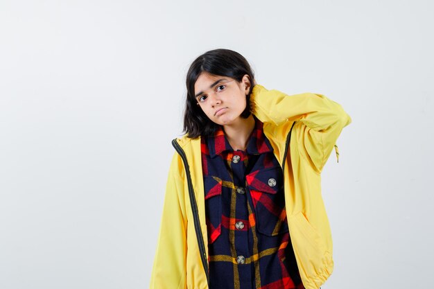 Little girl in checked shirt, jacket holding hand on neck and looking fatigued , front view.