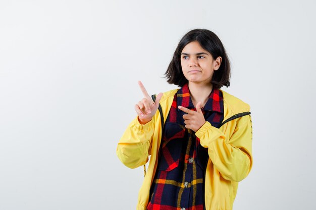 Little girl in checked shirt, jacket pointing aside and looking confident , front view.