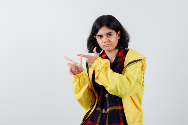 Little girl in checked shirt, jacket pointing to the left side and looking confident , front view.