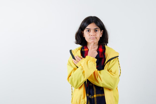 Little girl in checked shirt, jacket standing in thinking pose and looking thoughtful , front view.