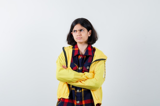 Little girl in checked shirt, jacket standing with crossed arms and looking gloomy , front view.