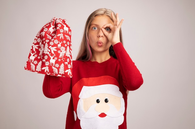 Little girl in christmas sweater holding santa red bag with gifts looking at camera surprised making ok sign over her eye standing over white background