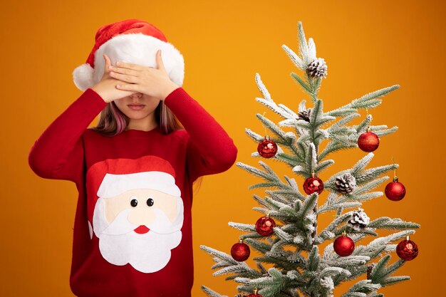 Little girl in christmas sweater and santa hat closing eyes with hands next to a christmas tree on orange background