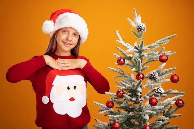 Little girl in christmas sweater and santa hat happy and surprised smiling cheerfully holding hands on her chest feeling thankful standing next to a christmas tree over orange background