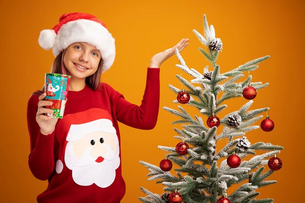 Little girl in christmas sweater and santa hat holding colorful paper cup presenting with arm of hand smiling standing next to a christmas tree over orange background