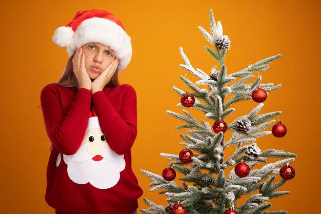 Маленькая девочка в рождественском свитере и новогодней шапке смотрит в камеру, смущенная и обеспокоенная, стоя рядом с елкой на оранжевом фоне