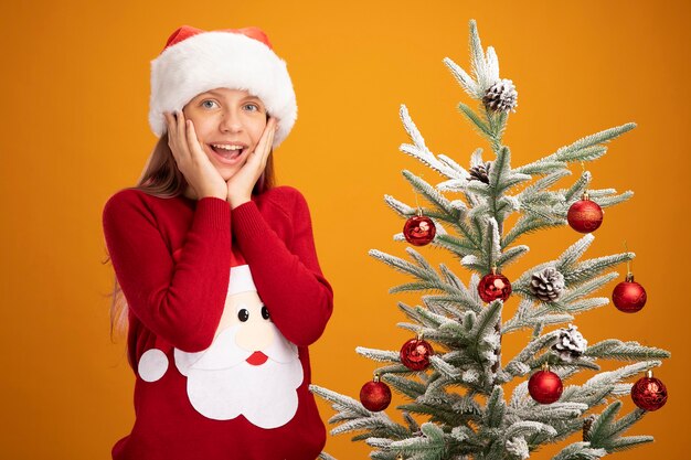 Маленькая девочка в рождественском свитере и новогодней шапке смотрит в камеру, счастливая и взволнованная, стоя рядом с елкой на оранжевом фоне