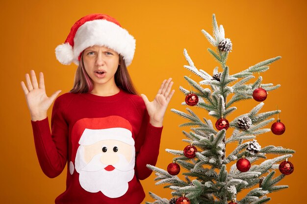 Маленькая девочка в рождественском свитере и шляпе санта-клауса смотрит в камеру, поднимая руки с разочарованным выражением лица, стоя рядом с елкой на оранжевом фоне