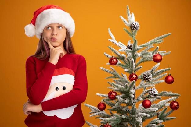 Little girl in christmas sweater and santa hat looking up with pensive expression next to a christmas tree on orange background