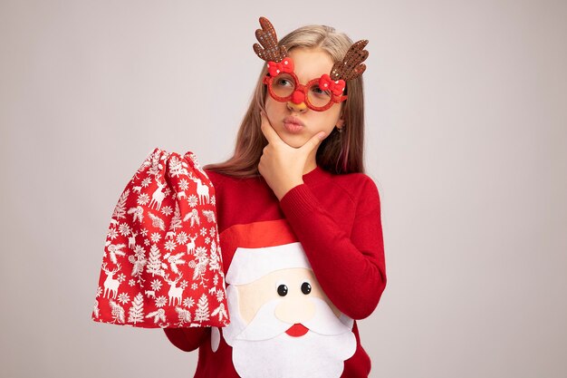 크리스마스 스웨터를 입고 크리스마스 스웨터에 어린 소녀 선물 산타 빨간 가방을 들고 옆으로 흰색 배경 위에 서 의아해