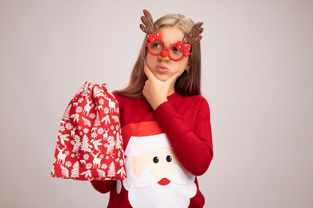 Little girl in christmas sweater wearing funny party glasses holding santa red bag with gifts looking aside puzzled standing over white background