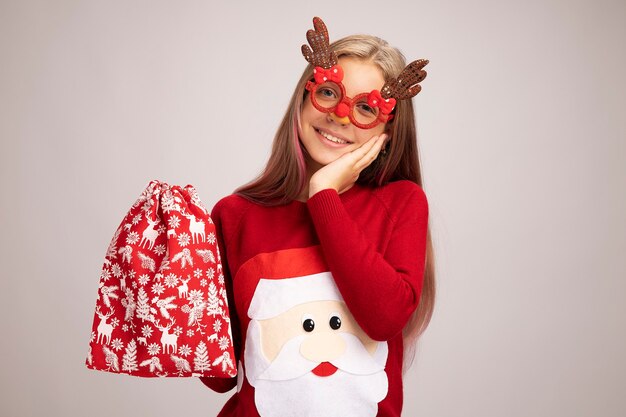 Little girl in christmas sweater wearing funny party glasses holding santa red bag with gifts looking at camera happy and positive smiling standing over white background