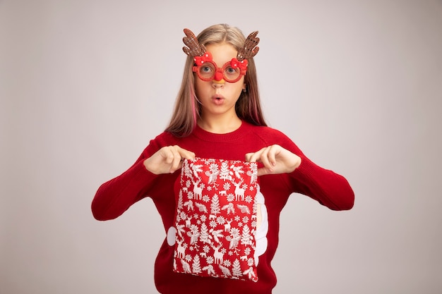 Little girl in christmas sweater wearing funny party glasses holding santa red bag with gifts looking at camera surprised standing over white background