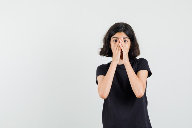 Little girl clasping hands on face in black t-shirt and looking scared , front view.