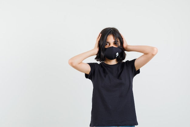 Little girl clasping head in hands in black t-shirt, mask and looking happy. front view.